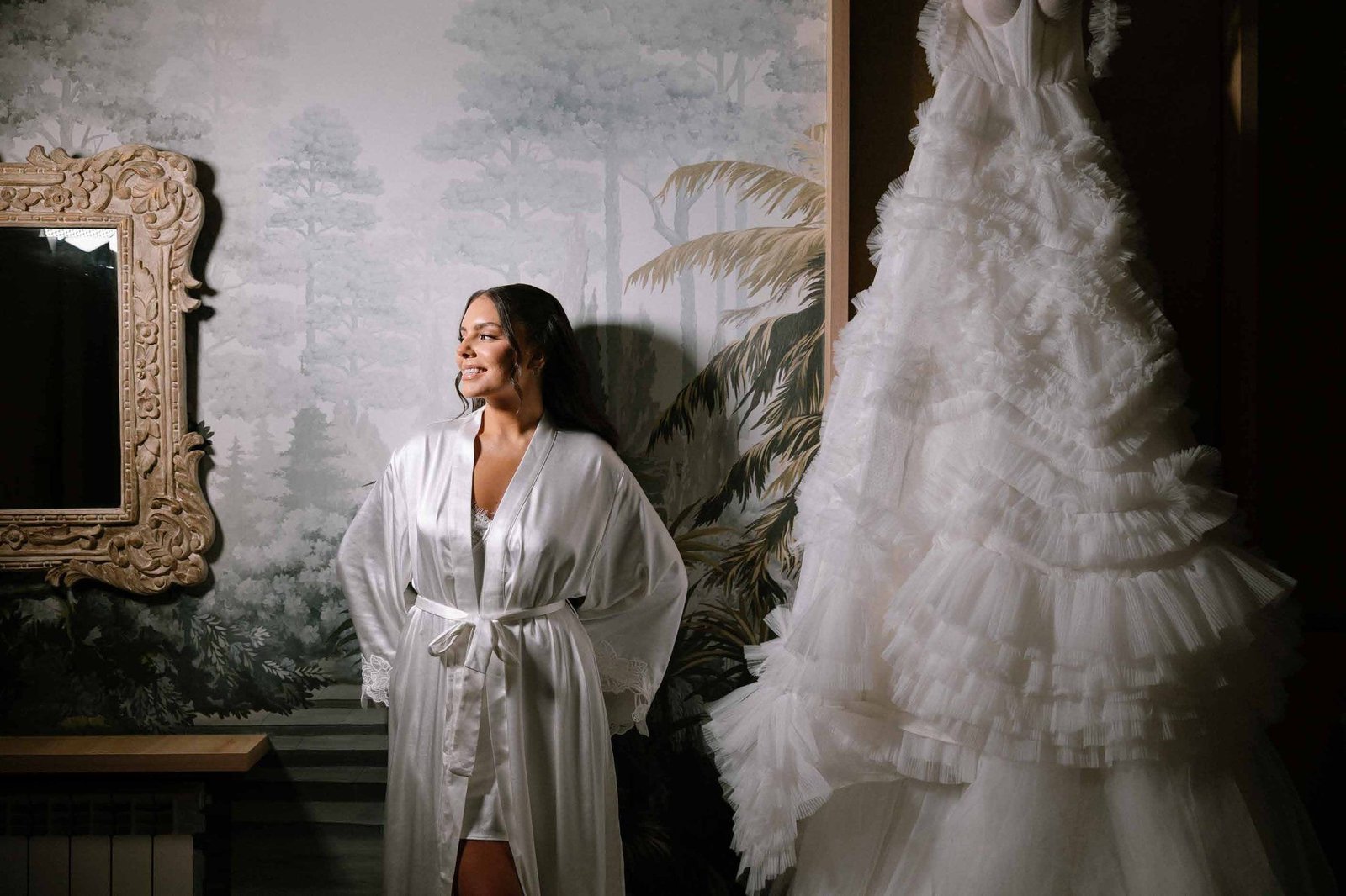 Bride in a silk robe beside her gown before a botanical mural